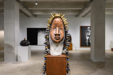 This image shows a gallery in The Andy Warhol Museum featuring a large, ornate mask sculpture with gold spikes and multicolored spherical decorations. It's centered in the room with abstract black-and-white artwork visible on the walls behind it. The gallery has a minimalist design with concrete floors and exposed beams.