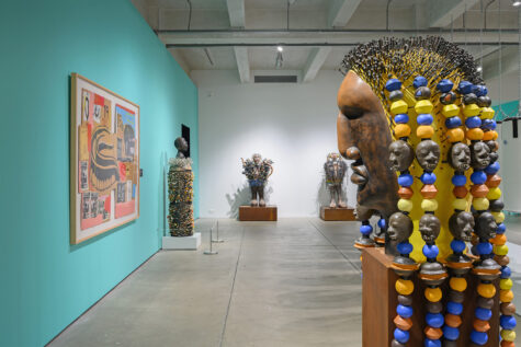 An art gallery at The Andy Warhol Museum featuring vibrant, eclectic sculptures and artwork. On the left, a wall displays a colorful mixed-media piece. In the center and right, several large sculptures with intricate designs made from various materials are exhibited. The sculptures include human-like figures adorned with colorful beads and textured surfaces, positioned under well-lit conditions. The gallery has a spacious, industrial feel with concrete floors and high ceilings.