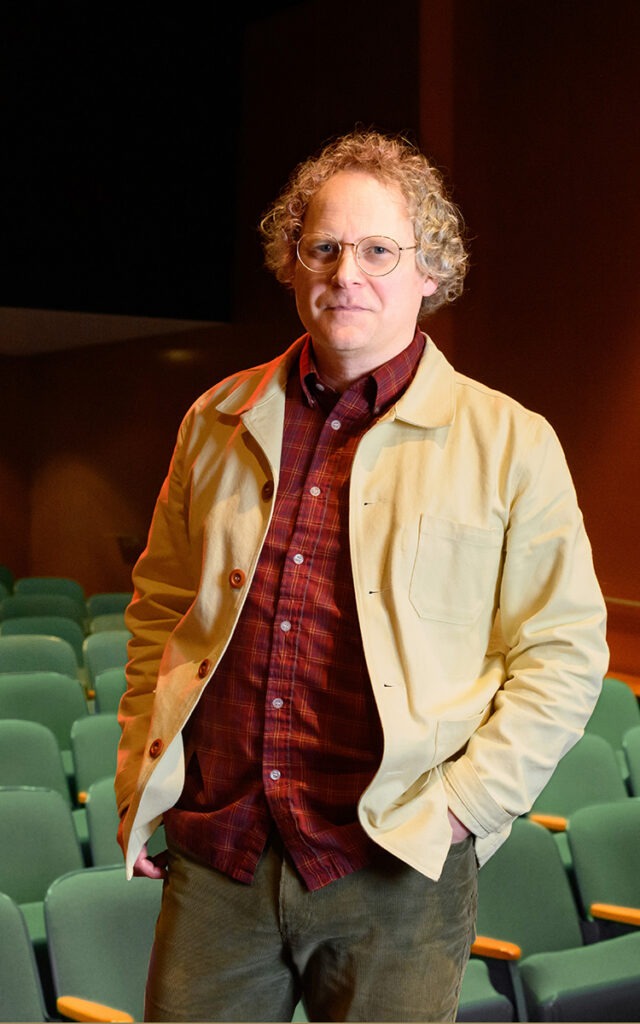 A person with curly hair and glasses stands in an auditorium wearing a beige jacket and red checkered shirt. Rows of green seats are visible in the background.