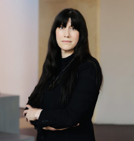 Hannah Klemm stands in a gallery space with white walls looking towards the camera. She has long, dark hair with bangs and is wearing a black shirt and black pants. Her arms are folded.
