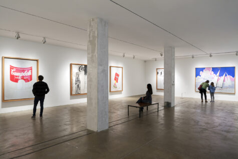 A spacious art gallery at The Andy Warhol Museum featuring several works by Andy Warhol, including a piece with a Campbell’s soup can theme. Visitors are observing the artworks; a couple stands close together, while another person sits on a bench. The room is well-lit, with white walls and a polished floor.